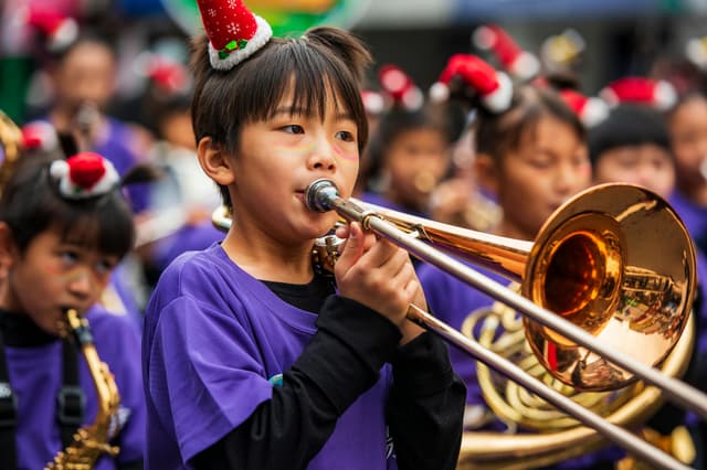 Child playing trombone in a festive parade, wearing a holiday hat, outdoors.