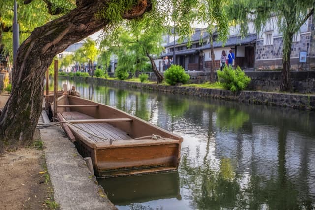 Scenic view of a traditional wooden boat docked along the canal in Kurashiki, Japan, surrounded by lush greenery.