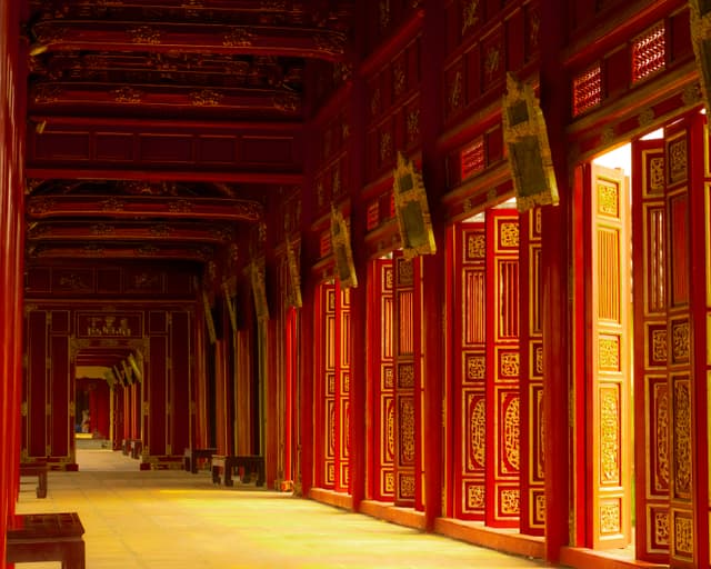 Intricate red corridor with open ornate wooden doors, featuring traditional architecture in warm sunlight.