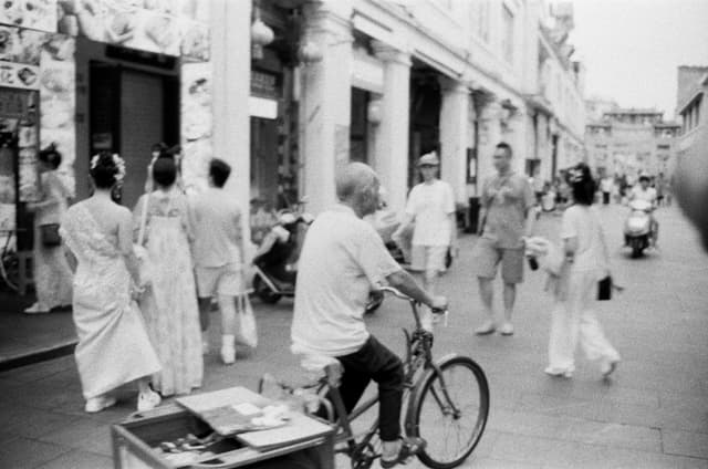 Black and white image of a busy street with people walking and a cyclist passing by.