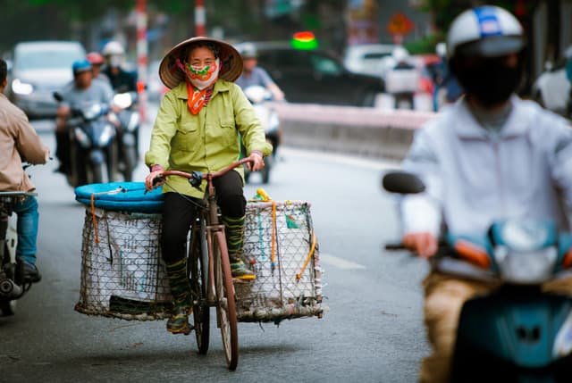 A woman cycling on a busy street in Hanoi, Vietnam, surrounded by motorbikes and wearing a traditional hat.