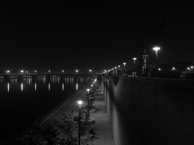 A stunning black and white night photo of a dimly lit riverside pathway with bright city lights reflecting on the water.