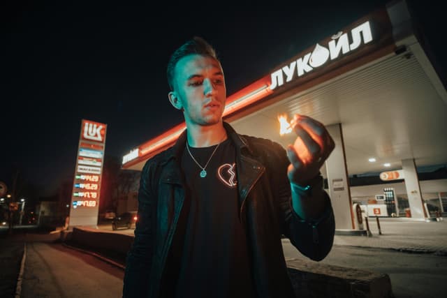 A man holding a lit matchstick stands in front of a Lukoil gas station at night, creating a dramatic scene.